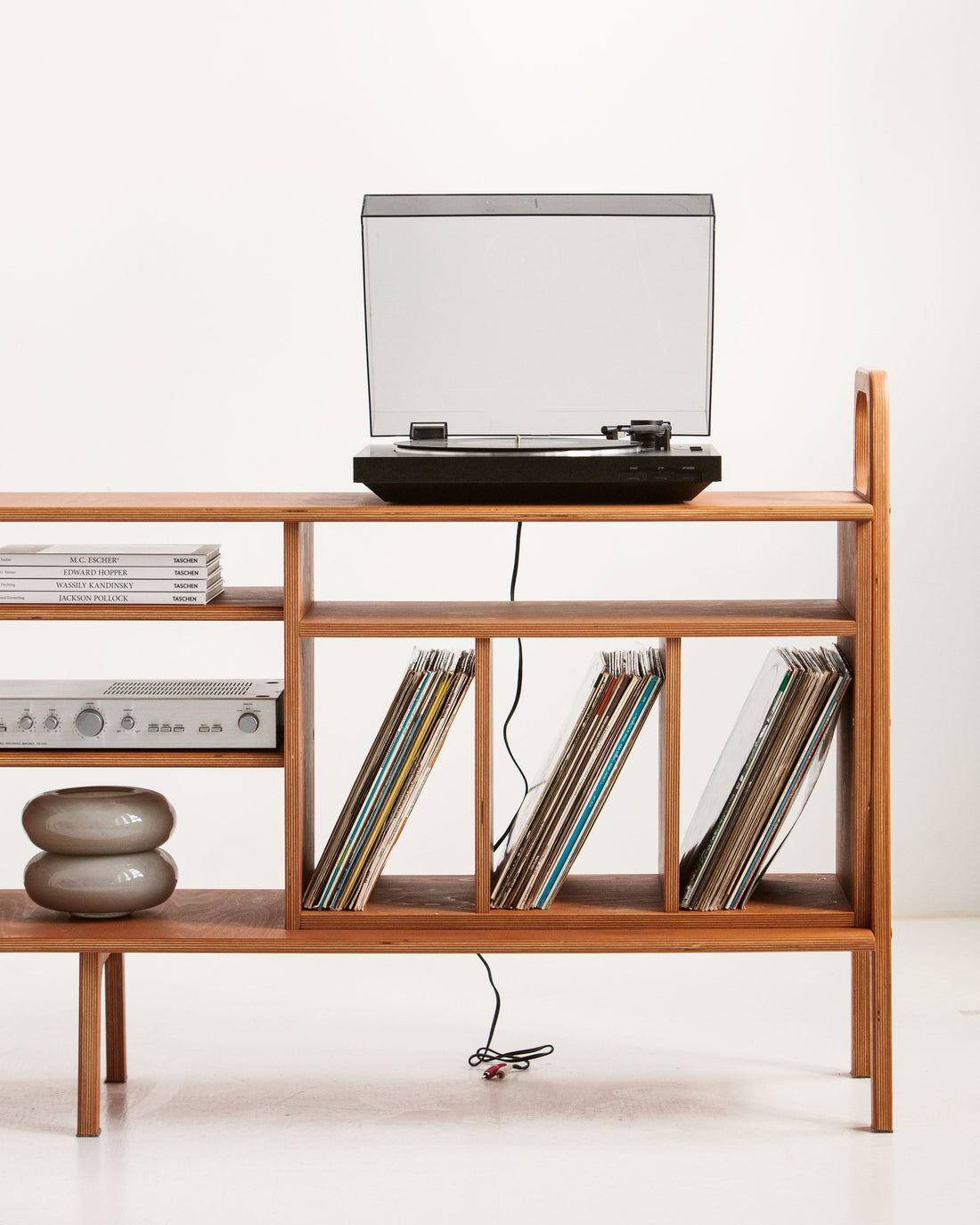 Turntable playing on a wide mid-century birch plywood vinyl record credenza, with an amplifier on the shelf below and 12-inch LPs stored upright in three divided bays