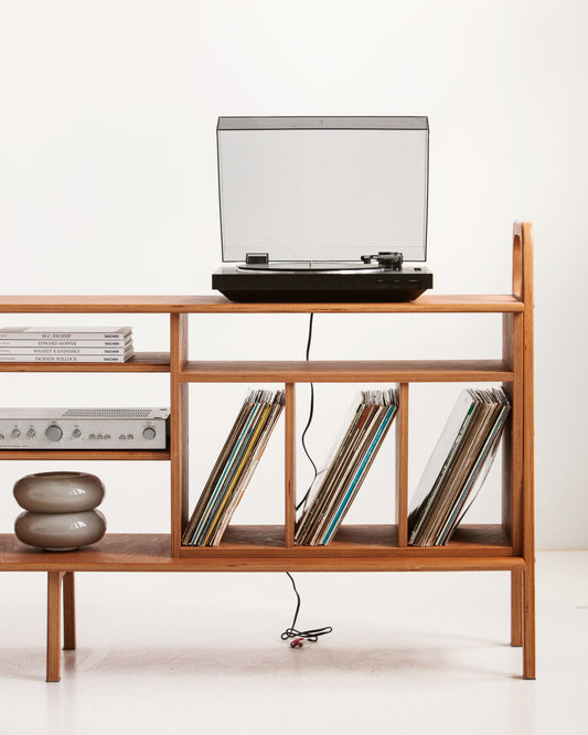Turntable playing on a wide mid-century birch plywood vinyl record credenza, with an amplifier on the shelf below and 12-inch LPs stored upright in three divided bays