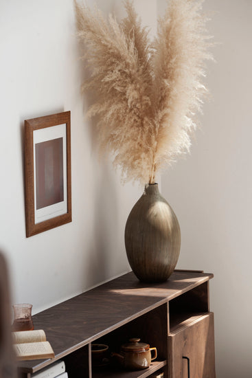 Decorative vase with dried pampas grass on a wooden shelf against a white wall.
